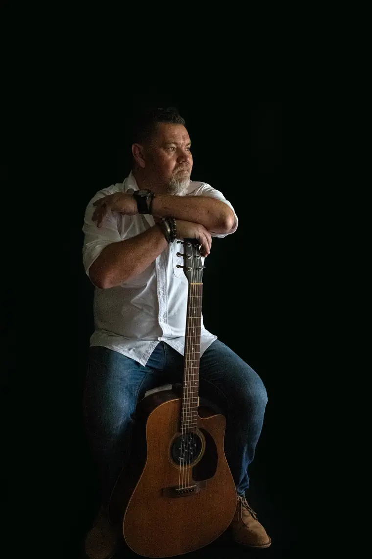 Wynand Strydom sit met sy kitaar oor die knie in ’n donker ateljee